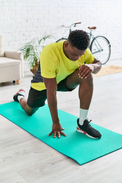 Atlético afroamericano hombre en ropa deportiva calentamiento en la estera de fitness en la sala de estar en casa - foto de stock