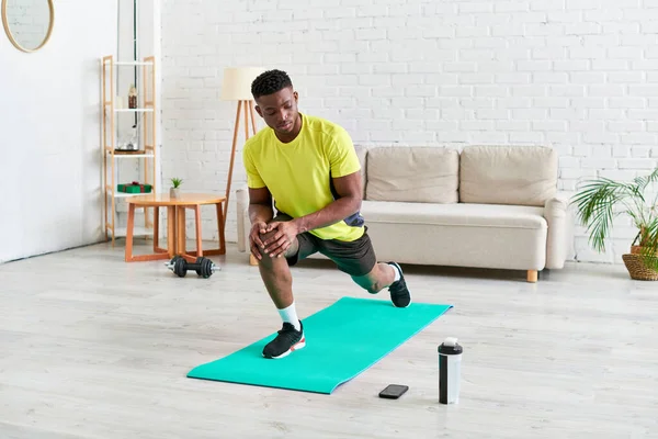 Desportivo afro-americano cara trabalhando fora em esteira de fitness perto de esportes garrafa e smartphone em casa — Fotografia de Stock
