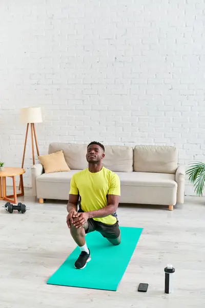 Joven afroamericano hombre haciendo ejercicio en la colchoneta de fitness cerca de botella de deportes y teléfono inteligente en casa - foto de stock