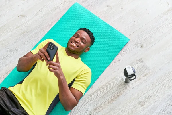 Hombre americano africano alegre que miente abajo en la estera de la aptitud con el teléfono inteligente cerca de la botella de los deportes, vista superior - foto de stock