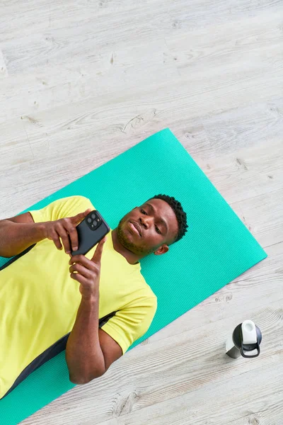 Joven afroamericano hombre acostado en la alfombra de fitness y navegar por Internet en el teléfono inteligente, vista superior - foto de stock