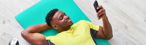 Alegre afroamericano chico acostado en la alfombra de fitness y navegar por Internet en el teléfono inteligente, pancarta - foto de stock