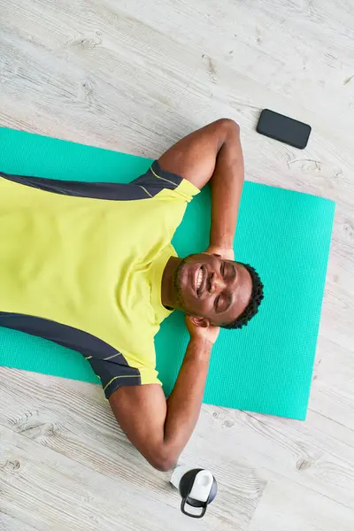 Vista superior del hombre afroamericano complacido en ropa deportiva acostado en la alfombra de fitness con los ojos cerrados - foto de stock