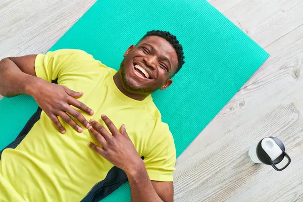 Hombre afroamericano feliz en ropa deportiva acostado en la alfombra de fitness y riendo con los ojos cerrados - foto de stock