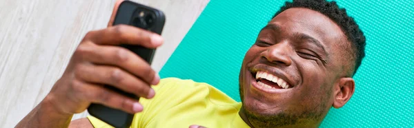 Riant homme afro-américain allongé sur tapis de fitness et naviguant sur les médias sociaux sur smartphone, bannière — Photo de stock