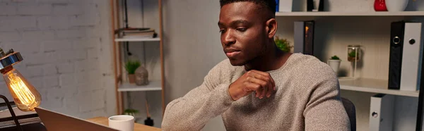 Jeune indépendant afro-américain concentré travaillant sur ordinateur portable le soir à la maison, bannière — Photo de stock