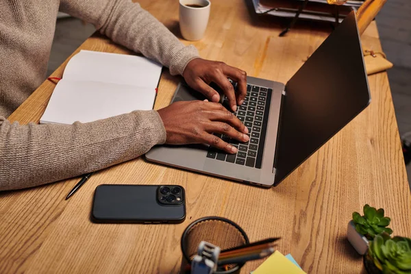 Vista recortada del hombre afroamericano escribiendo en el ordenador portátil mientras trabaja por la noche en casa, freelancer - foto de stock