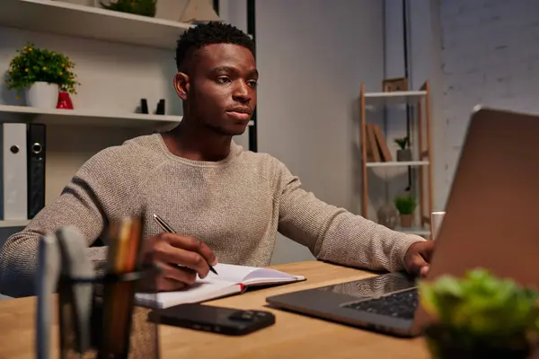 Afrikanisch-amerikanischer Freiberufler schaut auf Laptop und schreibt in Notizbuch, wenn er nachts von zu Hause aus arbeitet — Stockfoto