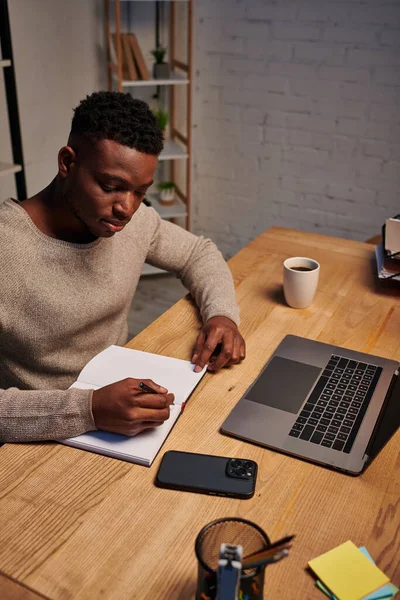 Afro-américain écrit dans un carnet près d'un ordinateur portable travaillant dans un bureau à domicile la nuit, pigiste — Photo de stock