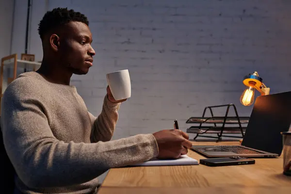 Afrikanisch-amerikanischer Typ mit Kaffeetasse schreibt in Notizbuch in der Nähe von Laptop, der nachts im Home Office arbeitet — Stockfoto