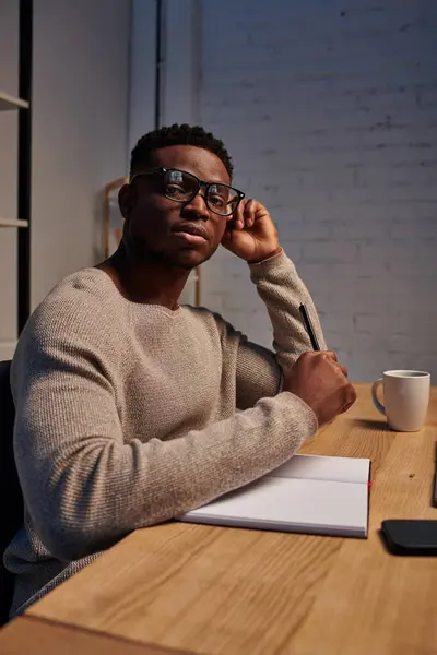 Афроамериканський чоловік в окулярах дивиться на камеру біля записника в домашньому офісі, фрілансер — стокове фото
