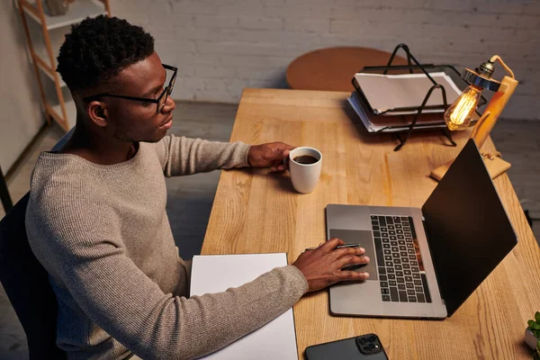 Афроамериканський чоловік в окулярах тримає чашку кави і працює на ноутбуці в домашньому офісі вночі — стокове фото