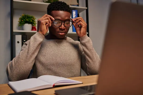 Young african american man adjusting eyeglasses near laptop and notebook while working at home — Stock Photo