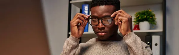 Joven afroamericano freelancer ajustando gafas mientras trabaja en casa por la noche, pancarta - foto de stock