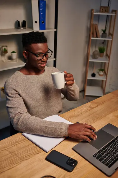 Усміхнений афроамериканський фрілансер в окулярах тримає чашку кави і працює на ноутбуці вдома — стокове фото