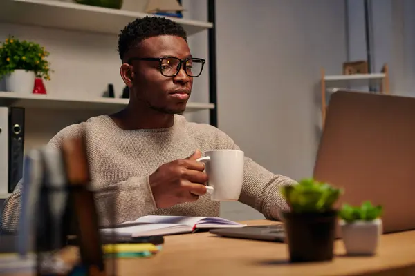 Зосереджений афроамериканський фрілансер в окулярах тримає чашку кави і працює на ноутбуці вдома — стокове фото