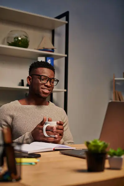 Complacido afroamericano freelancer en gafas con taza de café y mirando en el ordenador portátil en casa - foto de stock