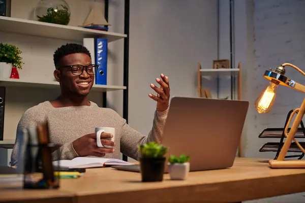 Счастливый африканский американец с чашкой кофе жестикулируя во время видеозвонка в домашнем офисе, фрилансер — стоковое фото