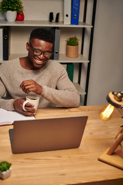 Веселий афроамериканський чоловік з чашкою кави сміється під час відеодзвінка в домашньому офісі, фрілансер — стокове фото