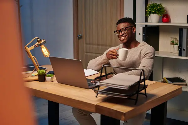 Радісний афроамериканський чоловік з чашкою кави посміхається під час відеодзвінка в домашньому офісі, фрілансер — стокове фото