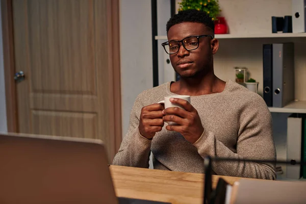 Hombre afroamericano reflexivo con taza de café mirando el ordenador portátil mientras trabaja en casa por la noche - foto de stock