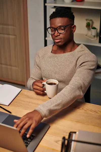 Hombre afroamericano concentrado con taza de café trabajando en el ordenador portátil en la oficina en casa por la noche - foto de stock