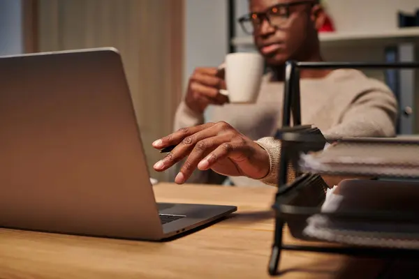 Giovane freelance afroamericano con tazza di caffè che lavora al computer portatile in ufficio a casa di notte — Foto stock