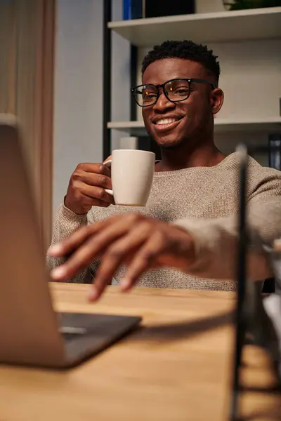 Радісний афроамериканський фрілансер з чашкою кави працює на ноутбуці в домашньому офісі вночі — стокове фото