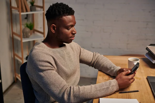 Guapo afroamericano freelancer navegar por Internet en el teléfono inteligente por la noche en casa, trabajo remoto - foto de stock