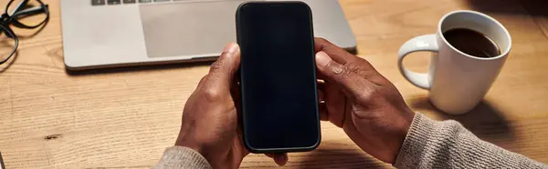 Vista recortada del hombre afroamericano sosteniendo teléfono inteligente con pantalla en blanco cerca de la computadora portátil, pancarta - foto de stock