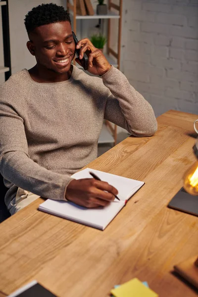 Fröhlicher afrikanisch-amerikanischer Mann, der während der Arbeit von zu Hause aus mit dem Smartphone in der Nähe von Laptop und Notebook spricht — Stockfoto