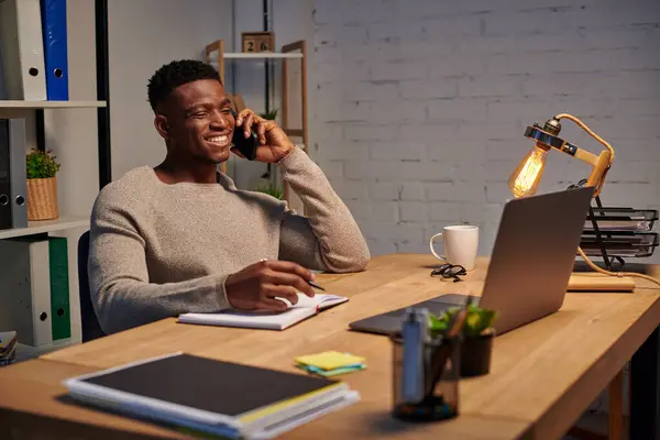 Homme afro-américain positif parler sur smartphone et écrire dans un carnet tout en travaillant à la maison — Photo de stock