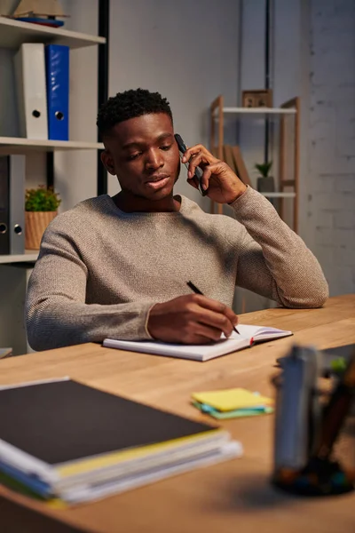 Serious african american freelancer talking on smartphone and writing in notebook, work from home — Stock Photo