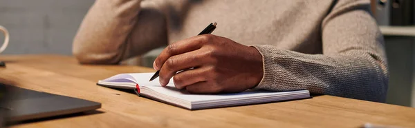 Vista recortada del freelancer afroamericano escribiendo en cuaderno mientras trabaja en casa, pancarta - foto de stock