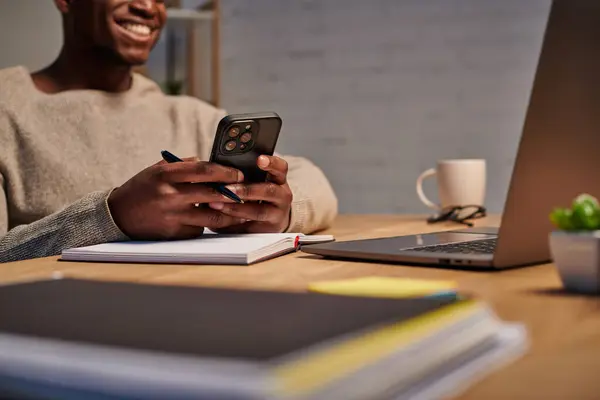 Усміхнений афроамериканський менеджер обміну повідомленнями на смартфоні біля ноутбука та ноутбука вдома, обрізаний вид — стокове фото
