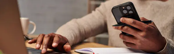 Vista recortada del hombre afroamericano con teléfono inteligente usando el ordenador portátil mientras trabaja en casa, pancarta - foto de stock