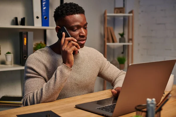 Freelancer afroamericano concentrado trabajando desde casa y hablando en smartphone cerca de laptop - foto de stock