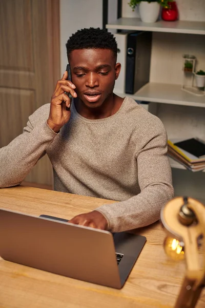 Junge fokussierte afrikanisch-amerikanische Freiberuflerin, die von zu Hause aus arbeitet und in der Nähe ihres Laptops mit dem Smartphone spricht — Stockfoto