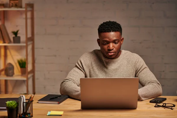 Молодий серйозний афроамериканський фрілансер, що друкує на ноутбуці вдома вночі, віддалена робота — стокове фото