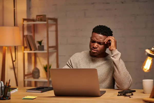 Réfléchi afro-américain pigiste regardant ordinateur portable dans le bureau à la maison la nuit, résolution de problèmes — Photo de stock