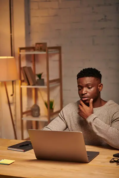 Pensativo afroamericano freelancer mirando el ordenador portátil en la oficina en casa por la noche, la resolución de problemas - foto de stock