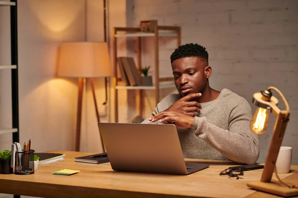 Thoughtful creative african american freelancer looking at laptop working from home at night — Stock Photo