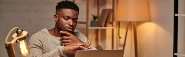 Creativo afro-americano freelancer pensare vicino laptop mentre si lavora da casa di notte, banner — Foto stock