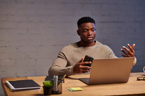 Молодий афроамериканський фрілансер з жестом смартфона під час відеодзвінка на ноутбук вдома — стокове фото