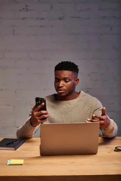 Serios afroamericanos freelancer con gestos de teléfonos inteligentes durante el chat de vídeo en el ordenador portátil en casa - foto de stock