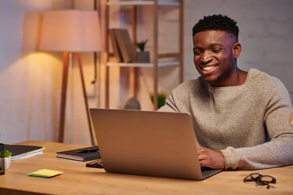 Веселий афроамериканський чоловік працює на ноутбуці вдома вночі, незалежний фрілансер — стокове фото