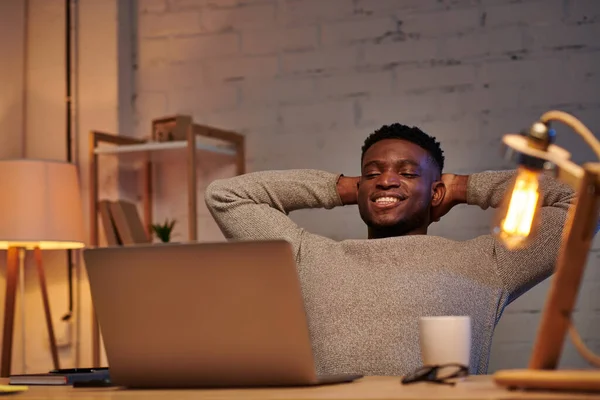 Piacere e rilassato uomo africano americano guardando il computer portatile in ufficio a casa di notte, libero professionista — Foto stock