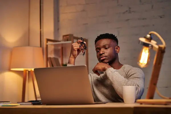 Riflessivo freelance africano americano che tiene occhiali da vista guardando il computer portatile in ufficio a casa di notte — Foto stock