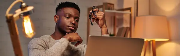 Pensativo afroamericano freelancer celebración de anteojos mirando portátil en casa oficina, pancarta - foto de stock