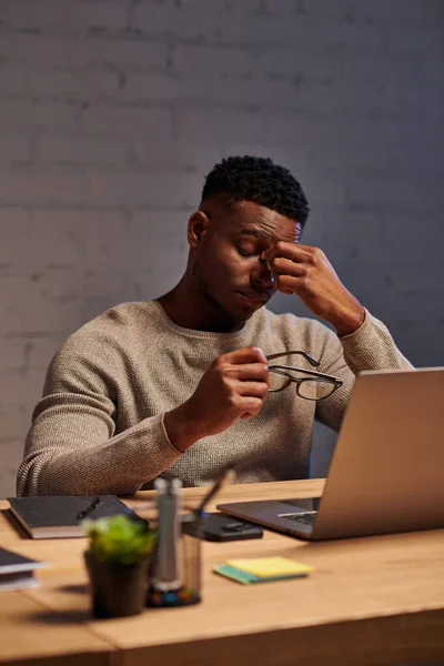 Esausto afro-americano freelance seduto con occhiali vicino laptop in home office di notte — Foto stock